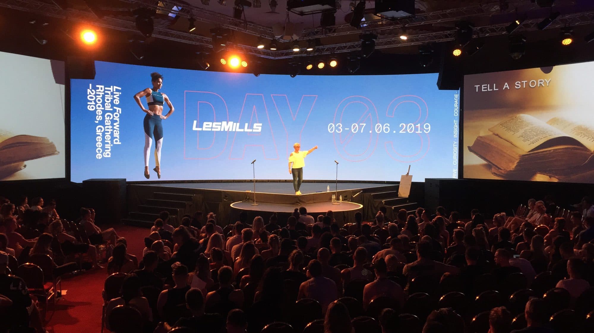A presenter on a wide stage at a packed fitness event